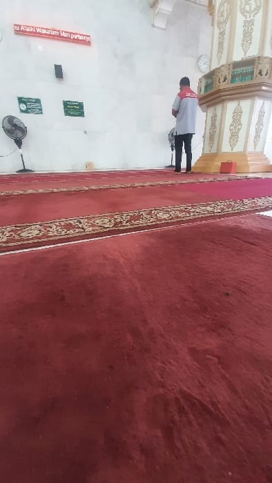 The image displays the interior of what appears to be a mosque or prayer hall, oriented as if viewed from a low angle, looking towards the back wall. The original image is rotated 90 degrees counter-clockwise.

The foreground and much of the floor space is covered by a plush, deep red carpet. The carpet features intricate patterned borders in a lighter gold or beige color, running along its length, suggesting lanes for prayer. The texture of the carpet is visible, showing some wear or nap variations.

In the middle ground, on the left side of the frame (when correctly oriented), a single person is visible, standing with their back to the camera. They are wearing a light-colored (possibly white or light grey) long-sleeved top with red sections on the shoulders/upper sleeves, and dark pants. They appear to be standing still, possibly in contemplation or waiting.

The walls are painted a light color, likely white or cream. On the far wall, to the left of the person, a round wall clock is mounted. Further to the left, below the clock, there are small, dark rectangular objects mounted on the wall, resembling speakers or other audio equipment. Near the bottom left corner of the image, a black electric fan stands on the floor.

Several signs are affixed to the wall. A prominent red banner or sign with white text is visible, partially obscured and difficult to read definitively due to blur and angle, but appears to say "tu Allahi Wasaallam Man parbaenye" or a similar religious phrase. Below this red sign, there are two smaller green rectangular signs with white text, which is too small and indistinct to decipher.

Architectural details are visible in the upper part of the background (when correctly oriented). These include decorative elements, possibly part of columns, arches, or wall panels, featuring elaborate gold or beige patterns against a light background. A small red box or container is situated on the floor in the background, near the decorative architectural elements.

The lighting suggests it is daytime, and the overall atmosphere appears quiet and serene, indicative of a place of worship.
