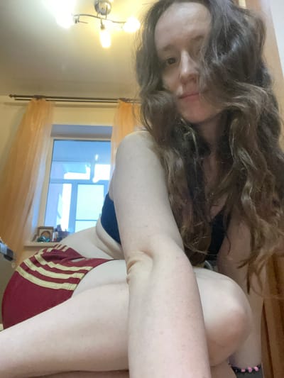 A person is in a room with a window and a sconce on the wall. The person has long brown curly hair. The person is wearing a blue sports bra and maroon shorts with gold stripes. The location cannot be identified as Usol'e, Russia, from the visual information provided.