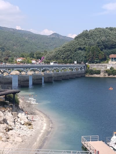 This is a daytime, vertical, eye-level shot of a dam and a body of water in Terras de Bouro, Portugal. The dam, constructed of concrete and stone, features a series of arches and supports, with a road on top where cars and a bus are visible. The water in front of the dam is a deep blue, with lighter, turquoise shallow areas near the shore. A sandy, rocky beach is visible on the left side of the frame, with several people swimming and sunbathing. A white boat is anchored in the shallow water near the beach. A wooden dock with white railings extends from the right side of the frame into the water. Mountains covered in dense green trees form the background, with a clear blue sky above dotted with white clouds. The scene depicts a recreational area by a dam on a sunny day.