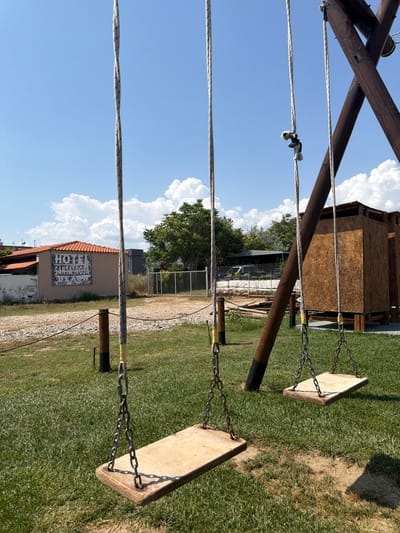 The image captures an outdoor scene on a bright, sunny day with a clear blue sky and fluffy white clouds, indicating midday or early afternoon due to strong shadows. In the foreground, two wooden swings hang from a rustic wooden swing set, attached by metal chains and ropes. The swings are empty, facing towards a grassy area and a gravel path.
In the mid-ground, a small wooden shed-like structure is visible, partially obscured by the swing set. Beyond this, a light-colored building with a terracotta tiled roof stands against the skyline. The building's side features a prominent mural composed of a grid of stylized letters and symbols. The word "HOTEL" is clearly visible in English on the upper right side of this mural. Other discernible elements within the grid include partial English words like "SANTA" and various Greek letters such as Lambda (Λ), Psi (Ψ), Theta (Θ), and Omega (Ω), suggesting a blend of languages, possibly for decorative or artistic effect.
Green grass covers much of the ground, interspersed with areas of dry earth and gravel paths. A tall tree with dense green foliage is visible to the right of the building, and a fence runs along the perimeter of the area. No people are present, depicting a quiet, recreational space in Alexandroupolis, Greece.