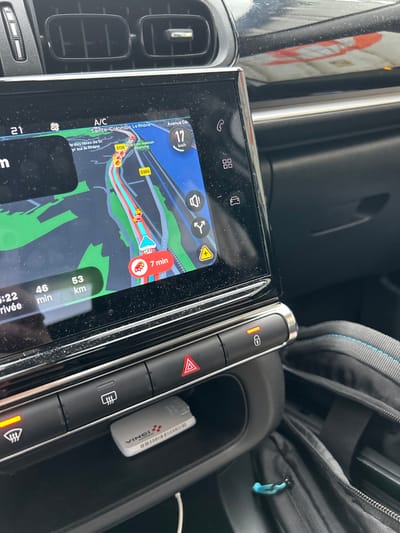 The image displays the interior of a vehicle, focusing on the central infotainment screen and a portion of the dashboard. The navigation system on the screen indicates the vehicle's current location on the D138 road in Saint-Cyr-sur-le-Rhône, France, with nearby textual references to Sainte-Colombe and Le Rhône, as well as Parcelle Maison Blanche and D386. The vehicle is moving at 17 km/h, with an estimated 46 minutes and 53 km remaining to the destination; a 7-minute delay is also indicated.
Below the screen, a row of control buttons is visible. The rear defroster button and the central door lock button are illuminated orange, signifying their active state. Beneath these controls, a small open compartment contains a white "VINCI Autoroutes" electronic toll transponder with a barcode visible on its side. Two horizontal air vents are positioned above the infotainment screen. A black bag, featuring blue stitching and a light blue zipper pull, is partially visible in the lower right corner of the frame. No individuals are depicted in the scene.
