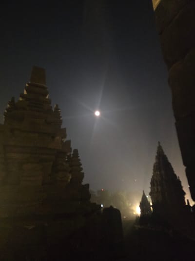 The image depicts a night scene in Bokoharjo, Indonesia. A bright moon dominates the sky. Silhouetted against the sky are the structures of the Plaosan Temple complex. The buildings appear to be made of stone. The lighting is dim. No people are visible in the scene.