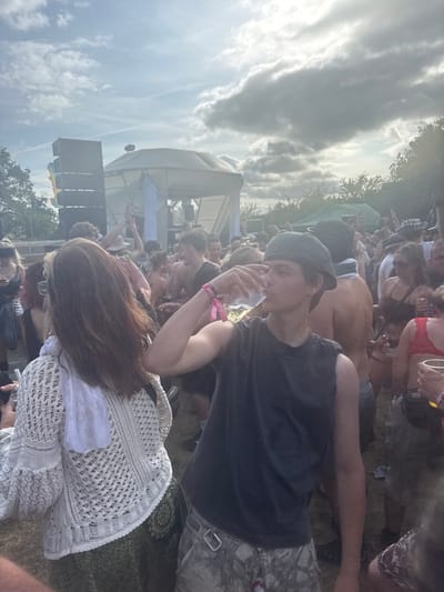 A sunny outdoor concert scene with a large crowd gathered near a stage. A young man in a dark shirt and cap holds a plastic cup, standing close to a woman with long brown hair wearing a white crochet sweater. Many other people are visible in the background, some shirtless. The cloudy sky suggests a bright, warm day.