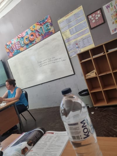 In the city of Dimovo, Bulgaria, a classroom scene unfolds. A woman, seated at a desk in the foreground, is engaged in reading a book. The room's walls are adorned with educational materials, including a colorful "WELCOME" sign and various grammar tables. A whiteboard displays handwritten text, possibly a quote or lesson. To the right, a wooden shelving unit holds books and supplies. In the foreground, a partially visible bottle of water rests on a table, with a book open beside it. The overall atmosphere suggests a learning environment, possibly during a break or before class begins.