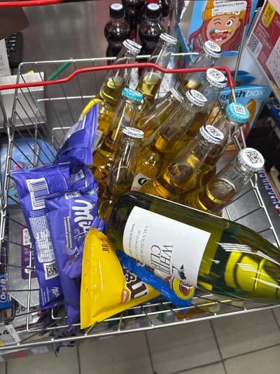 Captured from above, the image showcases a wire shopping basket filled with various items. Dominating the frame are multiple bottles of what appears to be light-colored beer, with caps bearing QR codes. A bottle of white wine labeled "White Cliff Sauvignon Blanc" is also present. Beneath these beverages, there's an assortment of confectionery items, including "Milka" chocolate bars, a yellow bag, and a box labeled "Freshbox Jelly Beans". The basket's red handle is visible on the left. The scene likely occurs in a retail environment in the city of Yemelyanovo, Russia.