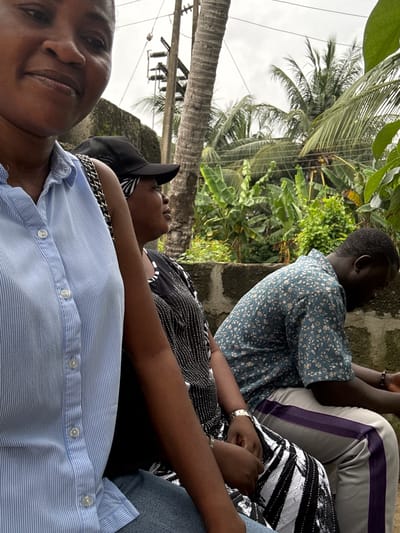 This is a vertically oriented photograph of four individuals, likely friends or family, seated together outdoors. The scene is lush and tropical, with prominent palm trees and other green foliage framing the subjects. The sky appears overcast, suggesting it might be a cloudy day or early morning/late afternoon.
In the foreground, a woman in a light blue pinstripe shirt is looking directly at the camera, her expression relaxed and her head tilted slightly. She is wearing a gold-toned chain necklace. To her left and slightly behind her, another person is seen wearing a black and white striped top with shiny embellishments. This individual has their arms resting on their lap.
Behind them, a person in a blue floral print shirt is partially visible, their back to the camera. They appear to be looking to the left, towards the foliage. Further back, a fourth individual is mostly obscured, wearing grey pants with purple and white stripes. Their legs are crossed, and they are also looking away from the camera.
The group is seated on what appears to be a low wall or ledge, possibly part of an outdoor seating area. The overall mood seems casual and relaxed, with the subjects enjoying each other's company in a natural, verdant setting. There is no visible text in the image.