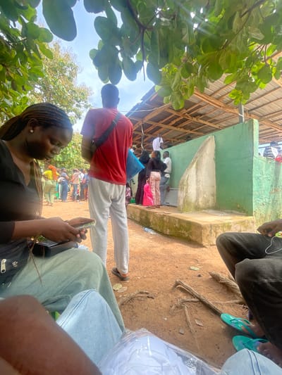 The image depicts an outdoor scene in Orun Ekiti, Nigeria, likely near a public facility. In the foreground, a woman with braided hair is seated and looking down at her mobile phone. To her right, a man in a red t-shirt and grey sweatpants stands facing away from the camera, with a dark strap across his chest. Behind them, a crowd of people are gathered, some appearing to be in a queue or waiting. The environment features a dirt ground, exposed tree roots, and overhead green foliage. In the background, there is a partially covered structure with a corrugated metal roof supported by wooden beams, and a green-painted wall with a concrete structure. Some individuals are standing near this wall, which has a water dispenser or fountain-like feature. The lighting suggests it is daytime, with sunlight filtering through the leaves.