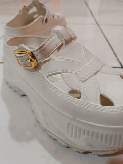 Two white platform sandals are placed on a tiled floor. The sandal in the foreground is a closed-toe shoe with a caged design on the upper and a strap with a gold buckle across the vamp. The platform sole has a ridged pattern. The other sandal is visible behind the first, appearing to be the same style. The environment suggests an indoor setting, likely a home, in Karangtengah, Indonesia, based on the information provided. The lighting is somewhat diffused, creating soft shadows.