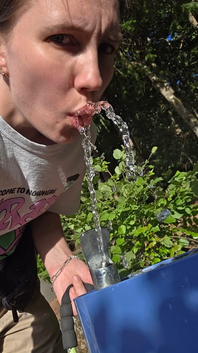A person is drinking water from a water fountain in Portland, Oregon. The person's face is visible from the nose up, and their mouth is open, receiving a stream of water. They are wearing a light gray t-shirt with text that reads "COME TO NOWHERE". A silver chain bracelet is on their left wrist. A blue container is positioned below the water fountain to catch excess water. The background consists of lush green foliage and trees. The lighting suggests it is daytime.