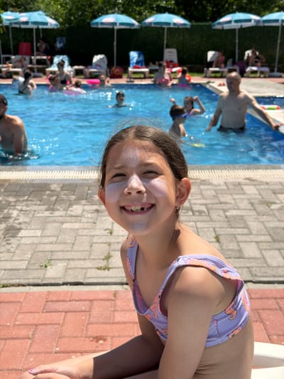 A young girl with a purple and pink patterned bikini is captured in a close-up, smiling with her eyes closed. She has white sunscreen on her cheeks and forehead. In the background, a swimming pool is visible, filled with people. Some are swimming, while others are relaxing by the poolside, under umbrellas. The scene appears to be a sunny day at an outdoor swimming pool, likely a public or resort pool, given the presence of lounge chairs and umbrellas. The paving stones around the pool suggest a well-maintained area. The overall mood is one of summer fun and relaxation. The orientation of the image is such that the girl is on the right side, and the pool extends to the left.