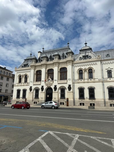 The image depicts the city hall of Craiova, Romania, from a ground-level perspective. The architectural style features ornate white facade with multiple windows, arches, and decorative elements. The sky above is partly cloudy. In the foreground, there is a street with marked parking spaces. A red car and a white car are parked along the side of the building. The street surface is gray, with visible yellow and blue markings. The image is taken from a slightly skewed angle.