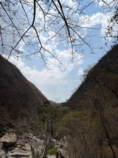 The image depicts a natural valley or canyon landscape in San Miguel Atlapulco, Mexico, under a bright, partly cloudy sky. The perspective is from an elevated position looking down into the valley. Steep, dry, vegetated hillsides frame the view, with the left slope appearing darker in shadow and the right slope illuminated. Dominant vegetation on the hillsides consists of deciduous trees and shrubs with minimal foliage, suggesting a dry season or climate. The foreground and midground feature a rocky terrain composed of large, light-gray boulders and smaller rocks, indicating a streambed or canyon floor.
Bare tree branches, largely devoid of leaves, are silhouetted against the sky in the upper portion of the frame. In the lower-mid section of the image, approximately ten to fifteen individuals are gathered among the rocks. Two individuals are seated on a large rock in the immediate foreground, facing away from the viewer. Further back, a larger group of people is visible standing and sitting on and around the rocks, positioned in what appears to be a natural gathering area. Their attire suggests recreational activity. Lush green foliage is present deeper within the valley, contrasting with the drier vegetation on the slopes. No specific dynamic actions or discernible interactions among the individuals are evident beyond their collective presence in the rocky environment.