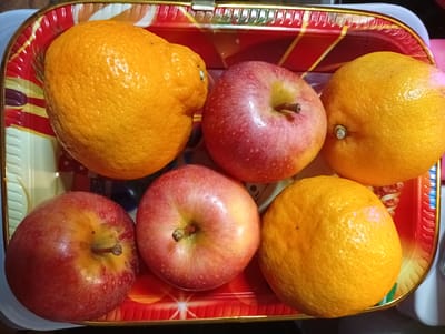 A rectangular red and gold serving tray holds six pieces of fruit: three round, red and yellow apples and three round, bright orange oranges. The tray is positioned on a flat, dark surface. The scene is evenly lit from above, casting soft shadows. The background is out of focus. The location is Ostrov-Zalit, Russia.