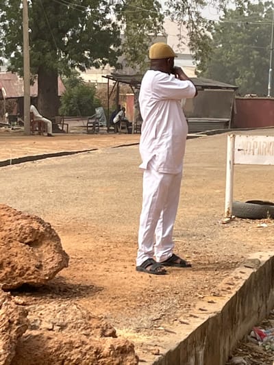 The image depicts an outdoor street scene in Kanti, Nigeria, under clear daylight conditions. In the mid-foreground, a man stands in profile, facing right, wearing a light-colored long-sleeved tunic over matching trousers and black open-toed sandals. He is also wearing a light brown flat cap, and his hands are raised towards his face. To his immediate right, a white sign post is visible, with "NO PARKING" text partially legible. An old tire rests on the dirt ground near the sign.
The ground consists of reddish-brown dirt, with a paved or compacted dirt path leading into the mid-distance. A concrete curb runs along the lower right edge, with some refuse visible beneath it. In the foreground to the left, a large, irregularly shaped brown rock or mound of earth is present.
In the background, beyond the path, several individuals and structures are visible. On the far left, a person sits on a white chair under a large tree with green foliage, positioned next to a utility pole. Further to the right, an open-sided shelter with a dark roof and support poles houses two or three indistinct figures. Other small buildings or shacks with corrugated metal roofs are scattered throughout the background, amidst additional green trees and foliage. The overall lighting suggests a bright, sunny day.