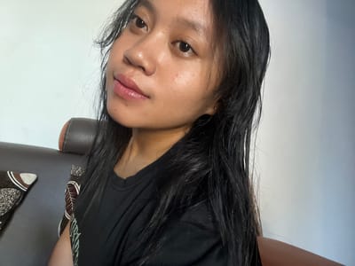 A young woman with long, dark hair is shown from the chest up, in a close-up shot, posing for a selfie indoors. She is wearing a black t-shirt. The background consists of a white wall and part of a brown leather sofa with patterned cushions visible on the left side of the frame. The lighting suggests the image was taken during the day, possibly near a window. The woman appears to be in a casual, home environment. The location is described as Jambearum, Indonesia.