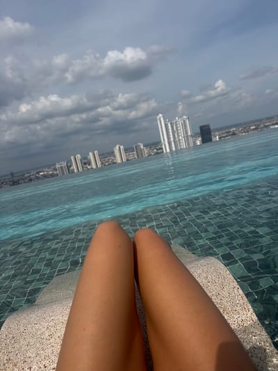 An overhead shot captures a person's tanned legs resting on a speckled white lounge chair in an infinity pool. The pool water is a clear turquoise with visible tiling below the surface. In the background, a cityscape unfolds with numerous tall, modern buildings under a cloudy sky. The location is identified as the city of Khan Chamkar Mon, Cambodia.