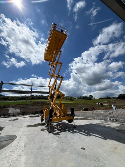 The image shows a bright yellow scissor lift, fully extended, positioned on a concrete surface at a construction site. The lift is in the foreground, angled towards the sky, with its platform at the top. In the background, there is a clear blue sky with scattered white clouds, and the sun is shining brightly, creating a lens flare effect in the upper left corner. Beyond the concrete area, there is a grassy hill with trees and some construction materials, including what appears to be metal beams and a portable toilet. The ground is a mixture of concrete, gravel, and some puddles of water. The overall impression is of an active construction site on a sunny day. The location is identified as Janowice, Poland.