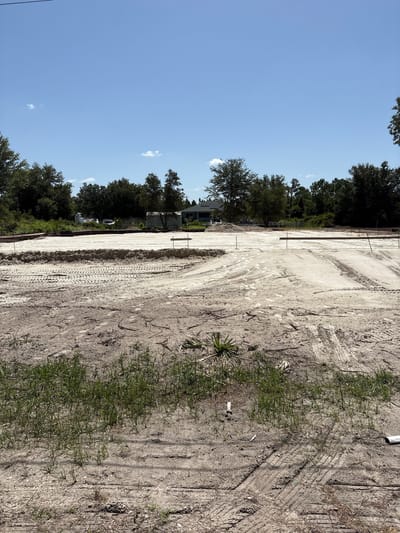 The image shows a construction site with freshly graded dirt and sand, featuring tire tracks from heavy machinery. In the background, a partially constructed building with a green roof is visible, surrounded by trees. The sky is clear and blue, with a few scattered clouds. The location is the city of Lehigh Acres, United States.