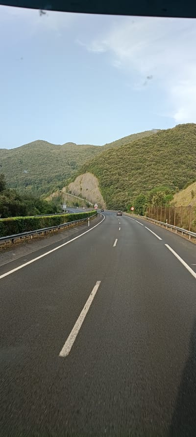 The image shows a view from a vehicle traveling on a highway through a mountainous, vegetated landscape. The highway is asphalt with white lane markings, and it curves to the right. On the left side of the road, there is a metal guardrail and a manicured hedge. The right side of the road is bordered by a fence and more vegetation. Steep, tree-covered hills rise on both sides of the highway. The sky is a light blue with some wispy clouds. There are a few cars visible on the road ahead, moving in the same direction. A speed limit sign is visible on the right side of the road. The overall impression is a scenic drive through a natural environment. It is not possible to determine the specific location in the city of Liendo, Spain, from the image alone.