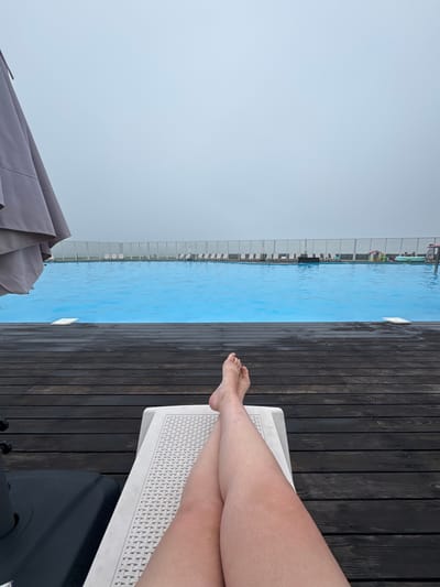 A person's legs are crossed and resting on a white lounge chair by an outdoor swimming pool. The pool has bright blue water and is surrounded by a wooden deck. In the background, there is a row of lounge chairs, a fence, and a cloudy sky. The overall scene suggests a relaxing day at a resort or hotel. There is no identifiable information about the city of Nord Ost, Russia in the image.