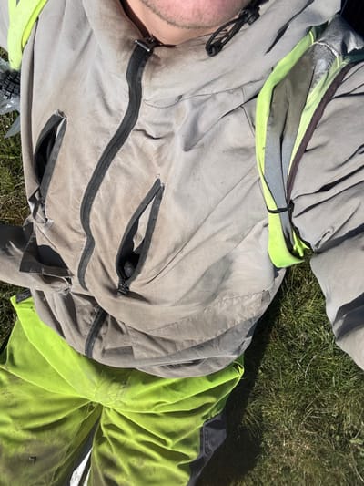 An overhead photograph depicts an individual from the mid-torso upwards, lying on green grass. The person wears a faded light grey jacket featuring a prominent central black zipper and two additional zippered pockets on the chest/abdominal area. The jacket appears soiled with dark stains. On the individual's right shoulder (left side of the image), a neon yellow-green high-visibility garment, containing grey reflective strips, is folded and secured with a black zip tie. The lower visible garment consists of neon yellow-green trousers, also soiled, with dark grey/black fabric panels towards the lower sections. The individual's chin and lower jaw are visible, exhibiting stubble. The environment is outdoors on green grass, illuminated by direct sunlight. The specific location within Southampton, United Kingdom, cannot be determined from the provided image.