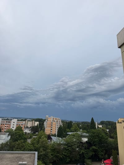 The image is an exterior, high-angle shot of an overcast sky above a dense urban and suburban landscape in Sindelfingen, Germany. In the foreground, the roof of a building is visible, made of grey material. The middle ground is filled with numerous trees and several multi-story residential buildings with brick and light-colored facades. One building in the center is predominantly orange brick with white accents. To the right, the corner of a yellow building is visible. In the distance, the landscape appears flatter with more trees and some faint structures under a dark, cloudy sky. The clouds are layered, with darker, more ominous clouds towards the bottom and lighter, more diffused clouds higher up. There is a single small object, possibly a bird or a drone, visible in the sky. No people are discernible in the image.