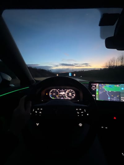 Inside a car, the dashboard glows in the dark. The steering wheel obscures a display screen, likely showing car diagnostics. To the right, a navigation system displays a map with route information. Outside, the sky transitions from dark blue to orange, indicative of dawn or dusk. The landscape includes trees and a signpost. The overall scene is a moment captured while driving.