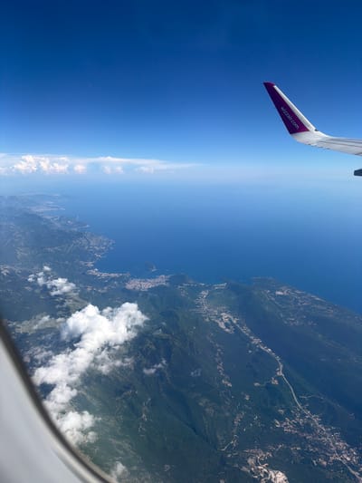 An aerial view from an airplane window shows a coastline with lush green mountains meeting the deep blue sea. A prominent airplane wing, with the Wizzair.com logo in purple, extends into the upper right corner of the frame. White clouds are scattered both above and below the aircraft, with a thin line of clouds visible on the horizon where the sky meets the sea. The coastline is dotted with small towns and settlements, with roads winding through the mountainous terrain and along the edge of the water. The city of Donja Cijevna, Montenegro, is visible along the coast, characterized by its dense development nestled between the mountains and the Adriatic Sea.