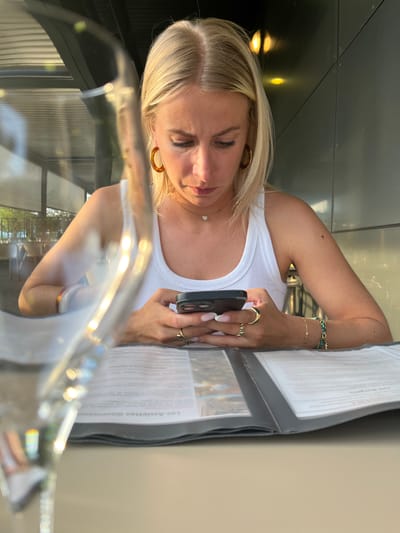 A blonde woman in a white tank top is seated at an outdoor cafe in Andrézieux-Bouthéon, France, looking at her phone. She is wearing gold jewelry, including earrings, rings, and bracelets. In front of her is a menu. The background shows a view of the city's outdoor scenery and buildings. A clear glass or plastic object is positioned in the foreground.