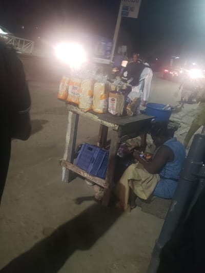 The image shows a street scene at night in Alalubosa, Nigeria. A woman is crouched near a wooden table on which several loaves of bread are stacked. She appears to be interacting with a mobile device, possibly making a transaction or communicating. A blue plastic crate sits underneath the table. To the left, a Mobil petrol station sign is visible, and cars are driving past. The scene is illuminated by streetlights and headlights, creating a high contrast.