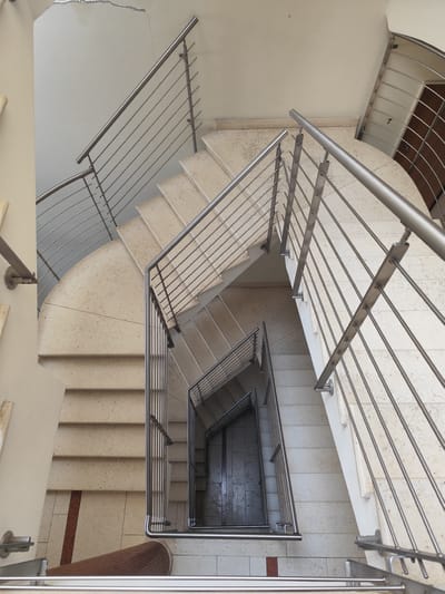 The image shows a top-down view of a modern, spiral staircase with light-colored stone steps and brushed metal railings. The staircase is located in a building in Spinea, Italy. The scene is indoors, and the lighting appears to be artificial, casting subtle shadows. There are no people or animals visible in the image. The focus is on the architectural details of the staircase, emphasizing its clean lines and contemporary design. The overall impression is one of a well-maintained, functional space.