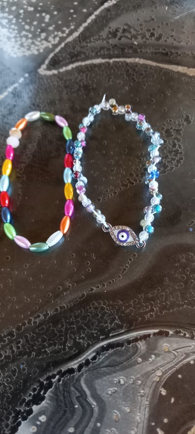Two bracelets are laid out on a dark, mottled surface. The bracelet on the left is made of colorful, oval beads in shades of pink, orange, white, green, blue, and red. The bracelet on the right is made of smaller, iridescent beads in clear, blue, pink, and gold. It also features a charm shaped like an eye, with a blue iris surrounded by silver rhinestones. The surface appears to be a table or countertop with a textured, almost abstract pattern. No people are visible in the image. The location is Iskra, Bulgaria, though this is not visually represented in the image itself.