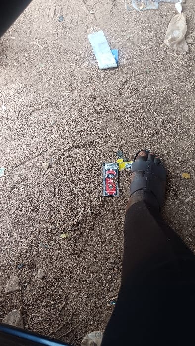 The scene depicts a high-angle view of an unpaved, dry ground surface, composed of sandy soil, fine gravel, and scattered debris. Resting on the ground near the center-right is a red and silver "KING" brand lighter featuring Chinese characters and a dragon design. Immediately adjacent to the lighter on the right is a dark-skinned left foot, partially visible and wearing a dark, multi-strapped open-toed sandal. In the upper left quadrant, a folded light blue plastic wrapper is present, with a crumpled, translucent plastic bag located further up and to its right. Various small fragments of discarded material are dispersed across the ground. A dark blue object, possibly a lower vehicle frame or structure, is partially visible along the bottom-left edge of the frame. The setting is outdoors under natural daylight, located in Langtang, Nigeria.
