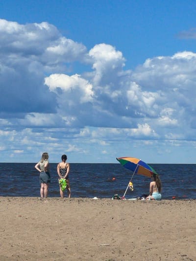 The image depicts a scenic view of a beach in Belozersk, Russia, on a sunny day with a bright blue sky and scattered white clouds. The foreground is a sandy beach, and the background features a large body of water, likely Lake Beloye, which is characteristic of Belozersk's location.
In the mid-ground, three individuals are present. Two women stand near the water's edge, facing away from the camera. The woman on the left wears a light-colored sleeveless top and dark shorts. The woman to her right is in a patterned one-piece swimsuit and has a bright green sarong draped over her arm. Further to the right, under a colorful beach umbrella, a third person, appearing to be a woman, sits on the sand, also facing the water. She is wearing a light-colored bikini.
The umbrella is tilted, casting a shadow. Various items, possibly beach bags or towels, are scattered on the sand near the umbrella. The water appears dark and calm, with a few distant orange buoys visible. The overall atmosphere suggests a leisurely day at the beach.
