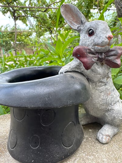 A grey, concrete garden statue of a rabbit wearing a dark red bow tie is positioned next to a black, top hat-shaped planter. The rabbit is leaning its front paws on the rim of the planter. The planter has several circular indentations around its base. Both objects are resting on a concrete surface. The background consists of lush green foliage and trees, suggesting an outdoor setting. The scene appears to be in Khazar Raion, Azerbaijan, given the context provided.
