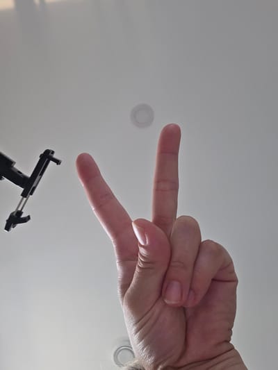 An overhead, low-angle shot shows a person's hand making a peace sign against a plain, light-colored ceiling. A black object, possibly a phone mount or tripod attachment, is visible in the upper left corner of the frame. The ceiling has a circular indentation or light fixture in the upper middle portion of the image. The lighting is diffuse and appears to be from an overhead source. The scene provides no information to determine its location in the city of Grünstadt, Germany.