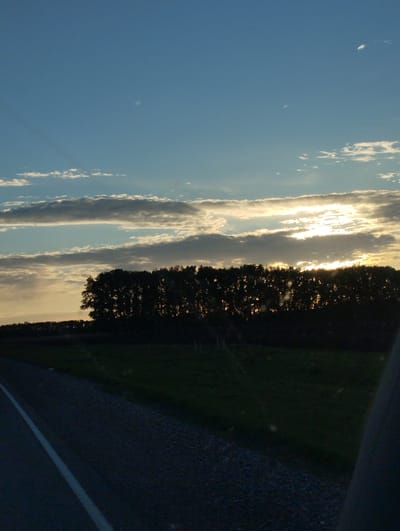 The image depicts a rural scene at dusk, not in Filippovo, Russia. The sky has a combination of blue and gray clouds with the sun setting behind a line of trees in the distance. The foreground shows a road with a white line marking the edge. There is a dark green field beyond the roadside. The image appears to have been taken from inside a vehicle.
