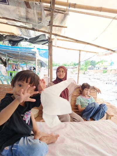 The image captures a candid scene in Meurah, Indonesia, under a makeshift, bamboo-framed shelter covered with various tarps, some of which are repurposed advertising banners (e.g., "TOP BRAND," "BIG DRE"). The lighting is bright, suggesting daytime.
In the immediate foreground, a young child with short dark hair, wearing a black t-shirt with a unicorn graphic and blue jeans, sits cross-legged on a light-patterned mat or cloth. The child faces the camera, holding both hands up in a motion that blurs them, with a crumpled white tissue or paper positioned near their right hand. The child's bare feet are visible.
In the midground, to the right of the foreground child, a woman with a maroon hijab and light brown top sits on a wooden slatted bench. She gazes to her left (viewer's right). Her left arm rests on the back of the bench. Beside her, another child with short dark hair, wearing a light green t-shirt with colorful patterns and blue jeans, also sits on the wooden bench, looking down and to their right.
In the background, under similar blue and translucent tarpaulin shelters, other individuals are visible. To the far left, people are seated at tables with green plastic chairs, some wearing hijabs. Beyond the immediate shelter structures, a wide, bright outdoor area is visible. This area appears to be a riverbed or a dry river, characterized by light-colored rocks and sandy patches, with some water visible. Multiple small figures of people are scattered across this expansive area, some near the water. On the distant bank, several white or light-colored buildings with red roofs are visible amidst green vegetation. The overall environment suggests a bustling, informal gathering or recreational spot alongside a river.