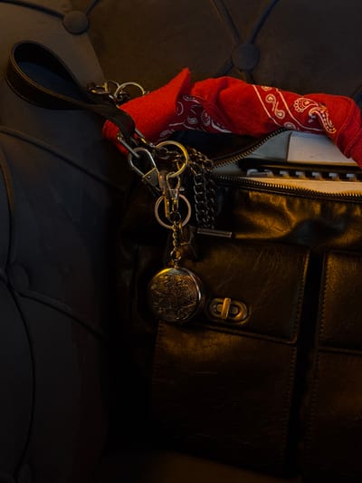 A dark, possibly black or very dark brown, textured leather or synthetic leather handbag rests on a dark, tufted upholstered surface, likely a chair or sofa. The bag features a main compartment with a partially open zipper revealing a folded red bandana with white paisley patterns, and beneath it, white papers or a spiral-bound notebook. Attached near the bag's dark strap are multiple silver-toned metal rings, carabiner clips, and chains, from which hangs a circular, antique-style metal locket or pocket watch adorned with an embossed floral or vine pattern. Two flap pockets are visible on the front of the bag, one secured by a brass-colored twist-lock clasp. The scene is dimly lit, with a warm, yellowish light source illuminating the right side of the bag and its visible contents, casting the left side and background into shadow. The location is Brčko, Bosnia and Herzegovina.