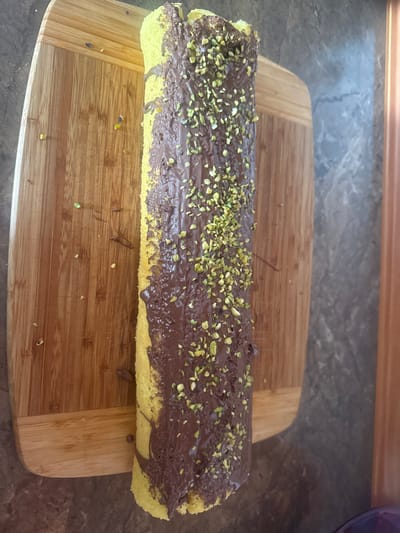 A close-up, top-down shot displays a rolled sponge cake covered in chocolate spread and sprinkled with chopped pistachios. The cake is placed on a wooden cutting board, which is situated on a patterned countertop. The lighting suggests an indoor environment, possibly a kitchen. The image does not provide any information about its location in Hraniv, Ukraine.