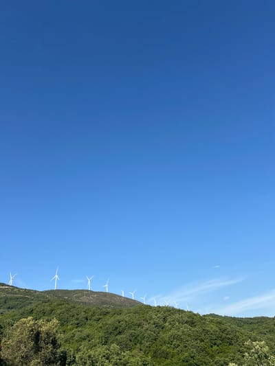 The image shows a clear blue sky over a hillside in Kruče-Kërruçi, Montenegro. Wind turbines are positioned along the ridge of the green, tree-covered hillside.