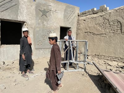 The image depicts an outdoor scene in Spin Boldak, Afghanistan, under a clear blue sky. Three young males are present amidst dusty ground and partially constructed or dilapidated buildings.
**People:**
1. **Foreground Left:** A young male in a dark tunic (shalwar kameez) and open-toed sandals stands facing slightly right. An orange fabric is draped over his left shoulder, and he wears a light-colored cap.
2. **Foreground Middle:** A young male in a brown tunic (shalwar kameez) and open-toed sandals walks towards the right, appearing in profile. He wears a light-colored cap and holds a bent metallic object in his right hand.
3. **Background Right:** A young male in a dark tunic (shalwar kameez) and a plaid shawl stands behind a metal frame, looking directly at the camera. He also wears a light-colored cap.
**Objects:**
* **Building Left:** A light gray, concrete or stucco-covered structure with an empty window opening on the left. Its facade features decorative raised elements: a five-pointed star, three diagonal lines, and three circular motifs with internal star-like designs. Cracks are visible in the surface.
* **Building Right:** An older, mud-brick structure, with significant portions of its outer rendering missing, exposing layers of mud bricks. The top edge of this wall appears uneven, suggesting damage or incomplete construction.
* **Metal Frame:** A disassembled or damaged light-colored metal frame, possibly from a window or gate, stands leaning against the right building. It contains fragmented glass or a similar clear material.
* **Fallen Object:** A large, flat, reddish-brown corrugated metal sheet or panel lies partially on its side on the ground to the right of the metal frame.
* **Ground:** The ground is unpaved, consisting of dry dirt, scattered rocks, and construction debris.
**Actions/Interactions:** The three individuals are standing in proximity within this outdoor space. The middle male carries a piece of debris, while the right male holds onto the metal frame. No direct interaction between the individuals is explicitly depicted; they appear to be observing their surroundings or the camera.