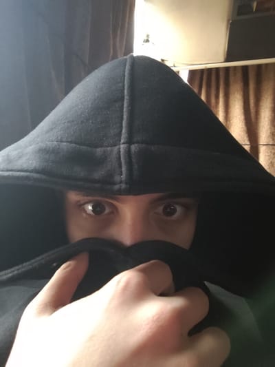 An indoor, close-up photograph taken in the city of Mankat al-Hatab, Syria, depicts an individual from the chest up. The subject wears a black hooded garment with the hood drawn over the head. The lower face, encompassing the nose and mouth, is intentionally obscured by the garment, held up by a light-skinned hand. Only the upper facial features are exposed; the eyes, dark brown and appearing dilated, are directed forward towards the camera. The immediate background features dark brown curtains on the left and a lighter brown, vertically textured curtain on the right. Above the right curtain, a section of a white ceiling and an overhead light fixture are visible. A portion of a green undergarment is discernible on the subject's right side, beneath the black hoodie.