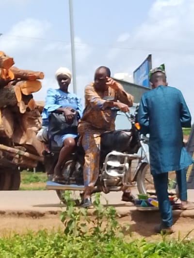 A man and two women are on a motorcycle loaded with large logs of wood. The man driving the motorcycle wears an orange and brown patterned shirt and pants. He is gesturing with his right hand. The woman behind him wears a blue dress and a white headscarf. She holds a dark bag on her lap. The third person, a man, stands to the right of the motorcycle, facing away from the camera. He wears a blue tunic and pants and a green and white patterned cap. The motorcycle is parked on a dirt road next to a patch of green foliage. In the background, there is a tall white pole and a building with a blue sign. The sky is cloudy. The scene appears to be outdoors during the day. The people are dressed in what appears to be traditional Nigerian clothing. The activity taking place is a pause in transport, with the driver interacting with the person to his right, possibly a vendor or acquaintance. The logs on the motorcycle suggest a transport of goods or materials.