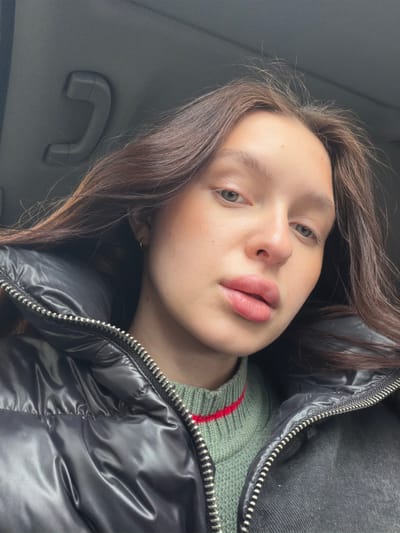 A close-up, slightly tilted portrait features a young person, likely female, with long, wavy brown hair framing her face. She has fair skin, prominent brows, and light-colored eyes, possibly green or hazel, looking directly at the camera with a neutral expression. Her lips appear full with a natural pink hue.
She is dressed in a shiny black puffer jacket, partially unzipped, revealing an olive-green knitted sweater underneath. A narrow red stripe is visible on the sweater's neckline or upper chest. The jacket's silver zipper teeth are a prominent detail.
The setting appears to be the interior of a vehicle, possibly a car, indicated by the dark gray textured ceiling and a visible gray grab handle positioned on the left side of the frame. The lighting is soft and even, suggesting an indoor or shaded daytime environment. The image is taken in a selfie style, capturing a casual and intimate moment. There is no visible text. The location context is Ilinskoe-Usovo, Russia.