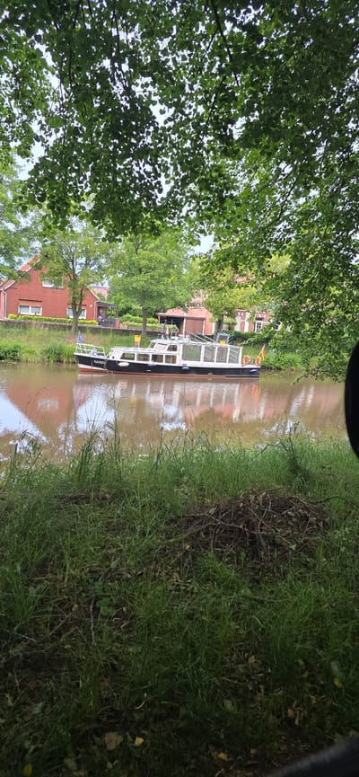 The image shows a canal in Haren (Ems), Germany, with a canal boat docked on the water. The boat is black and white with a glass-enclosed cabin. The canal bank is lined with green grass and trees, and red brick houses are visible in the background. The sky is overcast. The scene is tranquil and depicts a typical day in Haren (Ems).