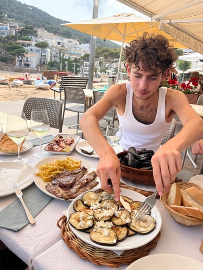 A young man with curly brown hair is sitting at an outdoor table, eating a plate of roasted eggplant and cheese.  He is wearing a white tank top.  He is using a fork to eat his food, and there are two other plates with food on the table, along with a glass of wine.  The table is covered with a white tablecloth.  The table is in front of a building with a view of the Mediterranean Sea.  The location is in Tamariu, Spain, which is located at the coordinates 41.918 latitude and 3.208 longitude.  The scene is a casual, relaxed dining setting.  The weather appears sunny.  The background is blurred with a building and sea in the distance.