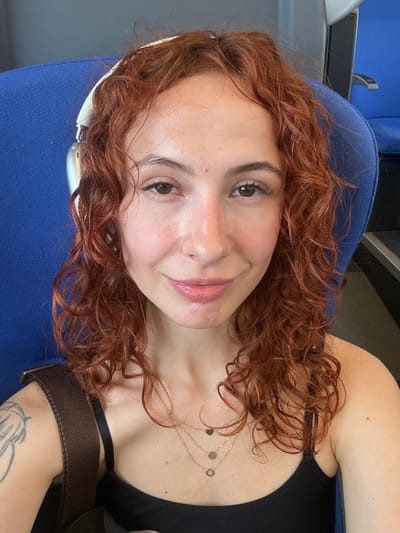 A young woman with reddish-brown, curly hair is captured in a selfie, smiling gently towards the camera. She is seated, likely in a public transport vehicle such as a train or bus, indicated by the blue upholstered seat and interior paneling visible in the background. The location context provided is Stara Pazova Municipality, Serbia.
She wears a black tank top or camisole and a layered gold necklace with small charms, including a heart, a circle, and a ring. White headphones are draped around her neck and ear. A brown strap, possibly from a bag, is visible across her left shoulder, and a line art tattoo depicting a stylized figure or design is on her left arm.
Her expression is relaxed and pleasant, with a slight smile. Her eyes are light brown, and she has light makeup, including mascara and a hint of lip gloss. The lighting suggests it is daytime. No visible text is present in the image.