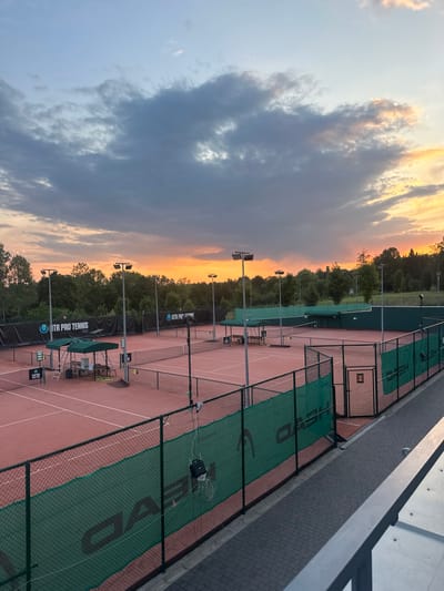The image displays an outdoor multi-court tennis facility located in Giebułtów, Poland, observed during sunset or dusk. The sky overhead transitions from a vibrant orange and yellow at the eastern horizon to lighter blues and grays with diffused clouds towards the zenith.
The scene features multiple red clay-surface tennis courts, each clearly delineated by white lines and equipped with a net. At least four full courts are discernible. The courts are enclosed by black chain-link fencing, to which green mesh banners displaying "HEAD" and "UTR PRO TENNIS" logos are affixed. Numerous tall metal poles supporting square or rectangular floodlights are positioned around and between the courts, indicating evening play capability.
Adjacent to some courts are green-canopied structures furnishing picnic tables and benches, providing seating or shelter. Elevated umpire chairs or scorekeeping stands are also visible near several nets. No individuals are present on the courts or within the immediate viewing area. A dark, rectangular bag is suspended from the green fencing in the lower-mid right section of the image.
In the foreground, a dark grey paver-stone walkway runs parallel to the court fencing, suggesting an elevated viewing or access path. Dense green deciduous trees and foliage form a natural perimeter around the entire tennis complex, extending into the background.