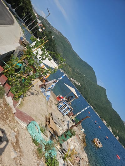This image depicts a coastal scene in Jošice, Montenegro, under a clear blue sky, suggesting a sunny day. The perspective is at an angle, tilting the view of the landscape. The foreground shows a rocky and gravelly area with some overgrown vegetation and a large coil of thick, turquoise rope. Red-tiled steps lead down from this area. To the left, a paved pathway curves away, with streetlights lining it.

In the middle ground, near the water's edge, there's a collection of objects, including stacked wooden pallets, a white tent-like structure with bamboo poles, some plastic bottles, and what appears to be a small kiosk or stand with a person visible underneath an umbrella. Further out in the tranquil, blue water, several orange buoys are scattered, along with a small, derelict boat.

The background is dominated by a steep, densely forested mountain that slopes down towards the water. Along the mountain's base, nestled amongst the trees, are clusters of white buildings, hinting at a town or village. The sea itself is calm, reflecting the bright sky. The overall atmosphere is one of a relaxed, seaside locale on a warm day, perhaps a less developed or working area of the coast, with elements of both natural beauty and human activity.